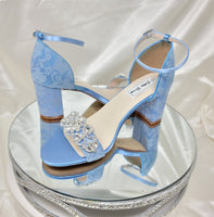 A pair of blue lace covered low block heel shoes with an ankle strap and a crystal design on the front toe strap of the shoes