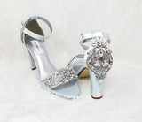 A pair of baby blue high block heel shoes with an ankle strap and a crystal and pearl design on the front toe strap of the shoes and a large crystal design on the back heel of the shoes