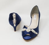 A pair of  bridal shoes in navy blue satin with a kitten heel and a peep toe and a crystal and pearl design on the front and back heel of the shoes