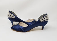 A pair of  bridal shoes in navy blue satin with a kitten heel and a peep toe and a crystal design on the front of the shoes and a crystal design on the back heel of the shoes