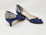 A pair of  bridal shoes in navy blue satin with a kitten heel and a peep toe and a crystal design on the front of the shoes and a crystal design on the back heel of the shoes