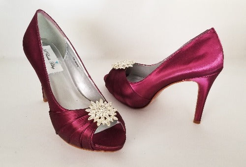 A pair of high heeled burgundy satin shoes with a peep toe and a hidden platform at the front of the shoes and a crystal design on the front of the shoes