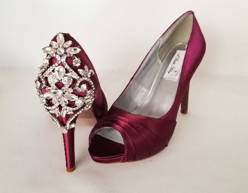 A pair of high heeled burgundy satin shoes with a peep toe and a hidden platform at the front of the shoes and a crystal design on the front of the shoes and a crystal design on the back heel of the shoes