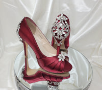 A pair of high heeled burgundy satin shoes with a peep toe and a hidden platform at the front of the shoes and a crystal design on the front of the shoes and a crystal design on the back heel of the shoes