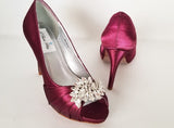 A pair of high heeled burgundy satin shoes with a peep toe and a hidden platform at the front of the shoes and a crystal design on the front of the shoes