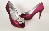A pair of high heeled burgundy satin shoes with a peep toe and a hidden platform at the front of the shoes and a crystal design on the front of the shoes