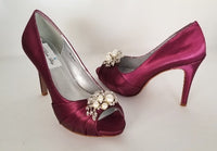 A pair of high heeled burgundy satin shoes with a peep toe and a hidden platform at the front of the shoes and a pearl and crystal design on the front of the shoes