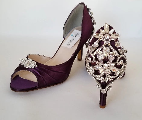 A pair of eggplant purple satin medium height heel bridal shoes with a peep toe and designed with a crystal design on the front of the shoes and a crystal design on the back heel of the shoes