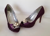 A pair of high heeled eggplant purple satin shoes with a peep toe and a hidden platform at the front of the shoes and a pearl crystal design on the front of the shoes