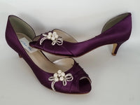 A pair of eggplant purple satin kitten heels with a peep toe and designed with a pearl and crystal bow design on the side of the shoes