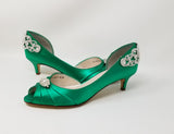 A pair of emerald green satin kitten heels with a peep toe and designed with a crystal design on the front of the shoes and a large crystal design on the back heel of the shoes