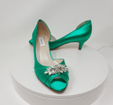 A pair of emerald green satin kitten heels with a peep toe and designed with a crystal design on the front of the shoes