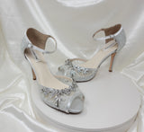 A pair of silver high heeled platform lace shoes with an ankle strap and a crystal design on the front of the peep toe shoes