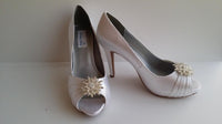 A pair of high heeled white satin shoes with a peep toe and a hidden platform at the front of the shoes and a crystal design on the front of the shoes