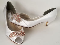 A pair of white satin bridal shoes with a kitten heel with a peep toe and a rose crystal design on the front of the shoes