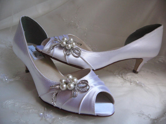 A pair of  white satin kitten heel shoes with a peep toe and a crystal and pearl design on the side of the shoes