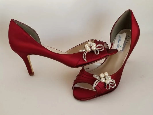 A pair of red satin medium height heel shoes with a peep toe and designed with a crystal and pearl bow design on the side of the shoes