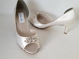 A pair of ivory satin medium height heel bridal shoes with a peep toe and designed with a crystal design on the front of the shoes