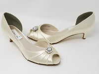 A pair of ivory satin kitten heel shoes with a peep toe with a small crystal design on the front of the shoes