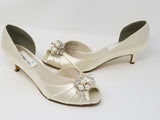 A pair of  bridal shoes in ivory satin with a kitten heel and a peep toe and a pearl and crystal design on the front of the shoes