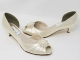 A pair of  bridal shoes in ivory satin with a kitten heel and a peep toe and a crystal design on the front of the shoes
