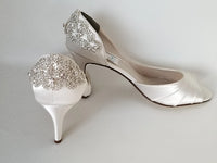 A pair of ivory satin medium height heel shoes with a peep toe and designed with a and crystal design on the front of the shoes and a crystal design on the back heel of the shoes