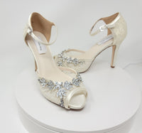 A pair of ivory high heeled platform lace shoes with an ankle strap and a crystal design on the front of the peep toe shoes