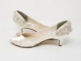 A pair of  bridal shoes in ivory satin with a kitten heel and a peep toe and a crystal design on the front and back heel of the shoes