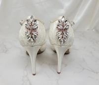 A pair of ivory high heeled platform lace shoes with an ankle strap and a crystal design on the back heel of the peep toe shoes