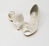 A pair of  bridal shoes in ivory satin with a kitten heel and a peep toe and a crystal design on the front and back heel of the shoes