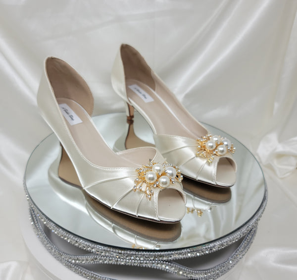 A pair of  bridal shoes in ivory satin with a kitten heel and a peep toe and a gold pearl and crystal design on the front of the shoes