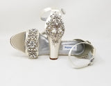 A pair of ivory high block heel shoes with an ankle strap and a crystal and pearl design on the front toe strap of the shoes and a large crystal design on the back heel of the shoes