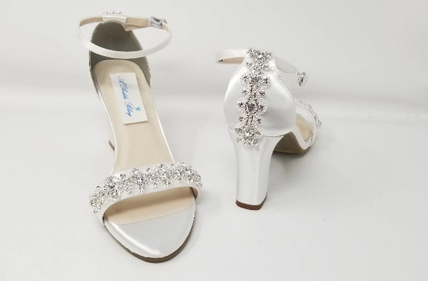 A pair of white block heel shoes with an ankle strap and a crystal design on the front toe strap of the shoes and a pearl and crystal design on the heel of the shoes