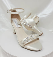 A pair of ivory block heel shoes with an ankle strap and a pearl and crystal design on the front toe strap of the shoes and a pearl and crystal design on the heel of the shoes