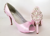 A pair of high heeled pink satin shoes with a peep toe and a hidden platform at the front of the shoes and a crystal design on the back heel of the shoes