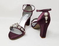 A pair of eggplant purple block heel shoes with an ankle strap and a crystal design on the front toe strap of the shoes and a crystal design on the heel of the shoes
