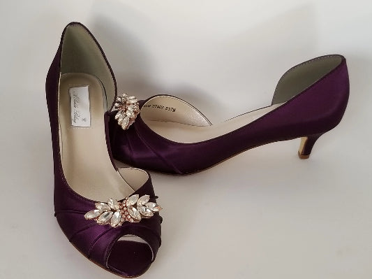A pair of eggplant purple satin kitten heels with a peep toe and designed with a rose gold crystal design on the front of the shoes