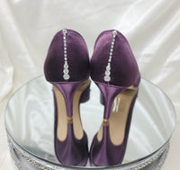 A pair of eggplant purple satin medium height heel bridal shoes with a peep toe and designed with a crystal design on the front of the shoes and a crystal teardrop design on the back heel of the shoes