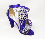 A pair of purple high block heel shoes with an ankle strap and a crystal and pearl design on the front toe strap of the shoes and a large crystal design on the back heel of the shoes