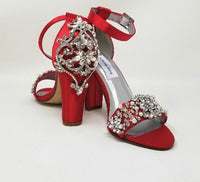 A pair of red high block heel shoes with an ankle strap and a crystal and pearl design on the front toe strap of the shoes and a large crystal design on the back heel of the shoes