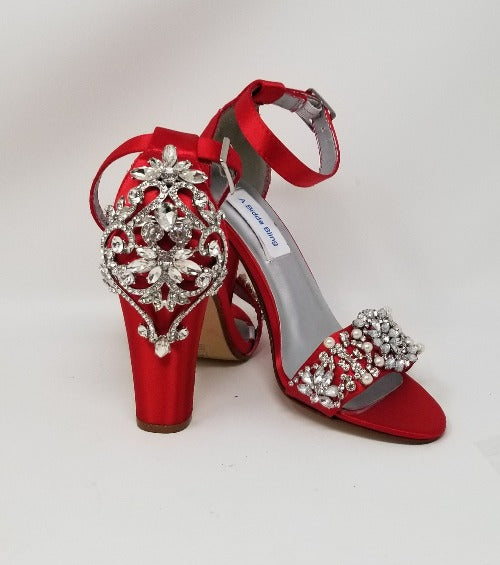 A pair of red high block heel shoes with an ankle strap and a crystal and pearl design on the front toe strap of the shoes and a large crystal design on the back heel of the shoes