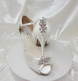 A pair of white high heeled platform lace shoes with an ankle strap and a rose gold crystal design on the front of the peep toe shoes and a rose gold design on the back heel of the shoes