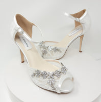 A pair of white high heeled platform lace shoes with an ankle strap and a crystal design on the front of the peep toe shoes