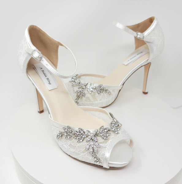 A pair of white high heeled platform lace shoes with an ankle strap and a crystal design on the front of the peep toe shoes