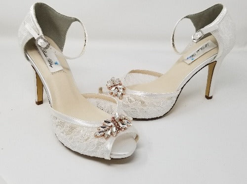 A pair of white high heeled platform lace shoes with an ankle strap and a rose gold crystal design on the front of the peep toe shoes
