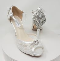 A pair of white high heeled platform lace shoes with an ankle strap and a crystal design on the front of the peep toe shoes and a crystal design on the back heel of the shoes