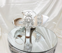 A pair of white block heel shoes with an ankle strap and a crystal design on the front toe strap and a crystal design on the back heel of the shoes