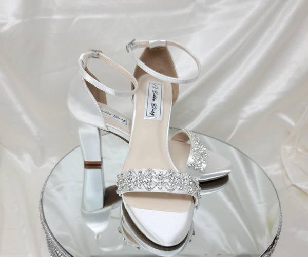 A pair of white block heel shoes with an ankle strap and a crystal design on the front toe strap of the shoes 