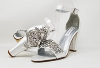 A pair of white high block heel shoes with an ankle strap and a crystal and pearl design on the front toe strap of the shoes and a large crystal design on the back heel of the shoes