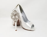 A pair of high heeled white satin shoes with a peep toe and a hidden platform at the front of the shoes and a crystal design on the back heel of the shoes